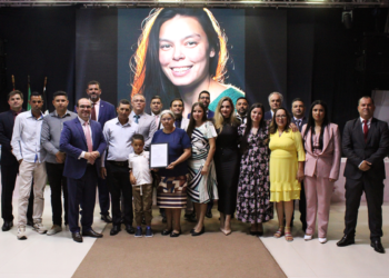 Autoridades, familiares e convidados posam em cerimônia na Câmara de Itararé em homenagem a Raquel de Oliveira Lima, com imagem da homenageada projetada ao fundo.