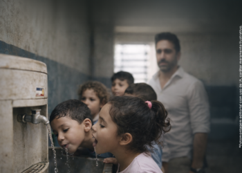 Crianças bebendo água em creche municipal de Itararé, com imagem desfocada do prefeito ao fundo, em ilustração editorial sobre falta de água potável.