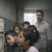 Crianças bebendo água em creche municipal de Itararé, com imagem desfocada do prefeito ao fundo, em ilustração editorial sobre falta de água potável.